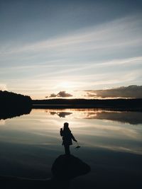 Silhouette child standing on rock against calm lake during sunset