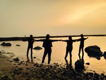 Rear view of friends with arms outstretched standing on shore at beach against sky during sunset