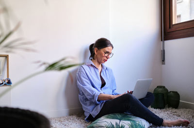 Concentrated barefoot female freelancer in eyeglasses typing on modern netbook while sitting on floor near white wall in light room