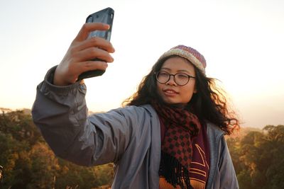 Woman taking selfie through mobile phone against sky during sunset