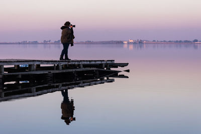 Woman photographing on pier over lake against sky during sunset