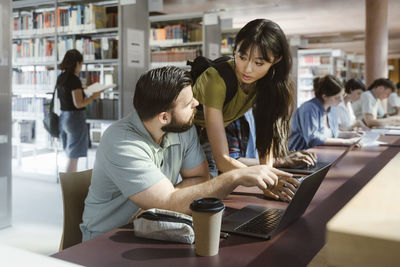 Young woman leaning near male friend talking while sitting at desk with laptop in university library