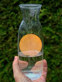 Cropped hand holding glass vase containing sun against green background 