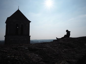 Man sitting on peak by old chapel against sky