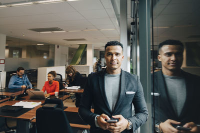 Portrait of confident mid adult businessman holding smart phone standing by reflection on window at office