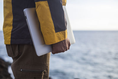 Midsection of man holding laptop with sea in background