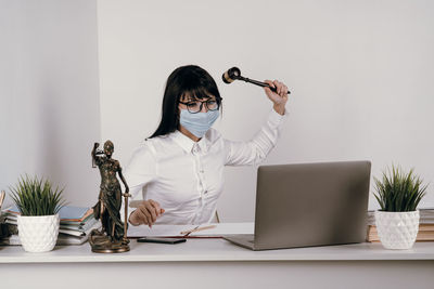 Young frustrated lawyer wearing mask hitting laptop by hammer at office