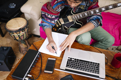 Young man with guitar writing notes on book by laptop in living room