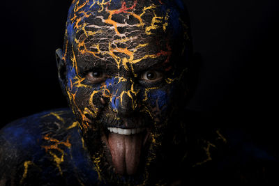 Close-up portrait of mid adult man with body paint sticking out tongue standing against black background