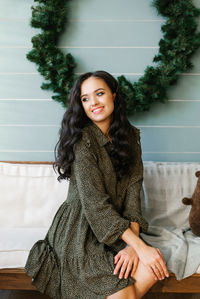 A young woman in a dress sitting on a light sofa against a blue wall with a christmas wreath 