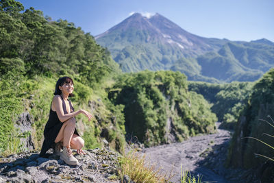 Full length of woman looking away while crouching on rock against mountains and sky