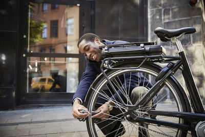 Smiling young male commuter locking electric bicycle while crouching against building in city