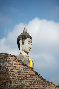 Low angle view of brick wall and buddha statue against cloudy sky