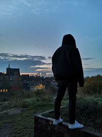 Rear view of man standing on retaining wall against sky during sunset