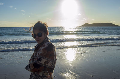 Portrait of young woman wearing sunglasses standing at beach against sky