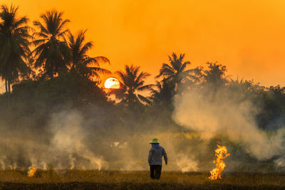 Rear view of man standing amidst smoky field against orange sky