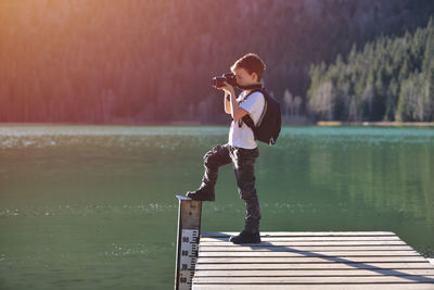 Boy photographing while standing on pier over lake