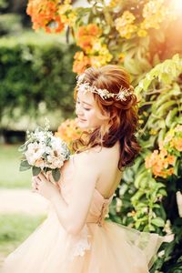 Side view of beautiful bride holding bouquet while standing by plants at park