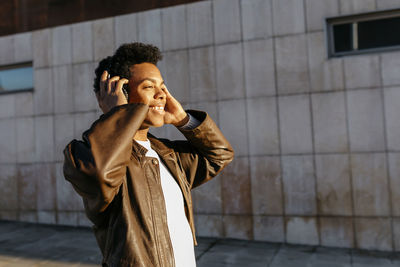 Smiling man listening music through wireless headphones against building during sunset