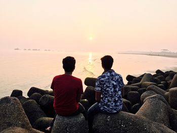 Rear view of friends sitting on rock by sea against clear sky