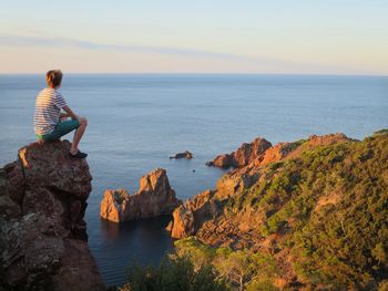 Rear view of young man sitting on cliff by sea