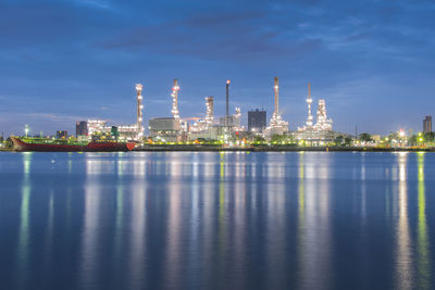 Illuminated factory by river against sky