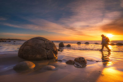 Man walking on beach by rock against orange sky