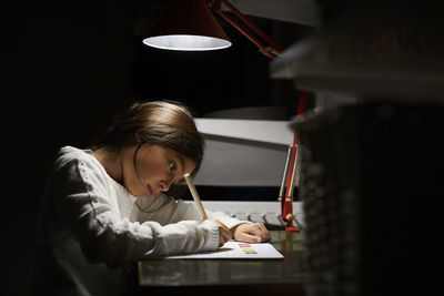 Girl studying while sitting at illuminated desk in darkroom