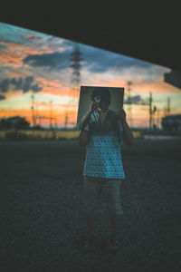 Rear view of person holding hat against sky during sunset