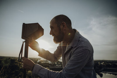 Side view of man looking through object during sunset against sky