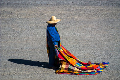 Side view of man wearing sombrero holding multi colored blanket while standing on street
