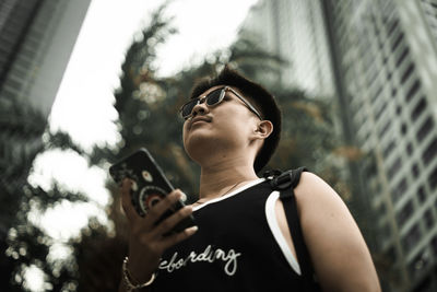 Low angle view of man wearing sunglasses while holding mobile phone against buildings