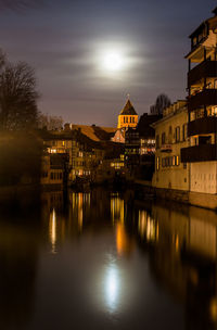 River by illuminated buildings against sky at sunset