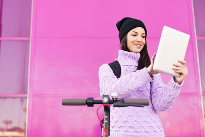 Young woman using digital tablet while standing against pink wall