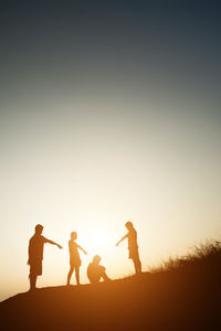 Silhouette people pointing over person sitting on cliff against sky during sunset