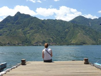 Rear view of mid adult woman sitting on pier over lake against mountain