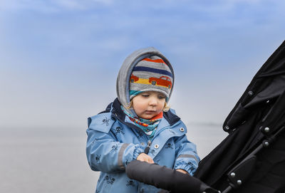 Cute girl wearing warm clothing while holding baby stroller against sky during winter