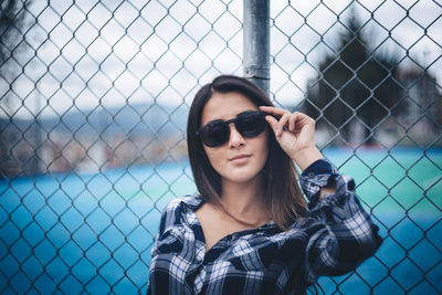Beautiful young woman wearing sunglasses leaning on chainlink fence