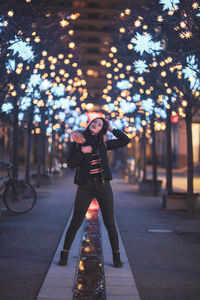 Portrait of woman standing on footpath against illuminated lights at night