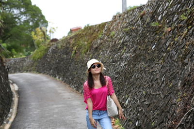 Young woman wearing sunglasses while walking on road by surrounding wall