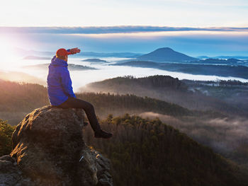 Hiker in blue jacket sit alone on the rock summit. heavy mist in deep valley. man shadowing eyes
