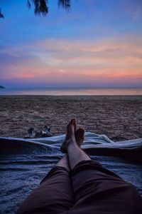 Low section of man relaxing on beach against sky during sunset