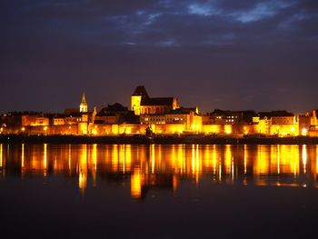 Illuminated buildings by lake against sky at night