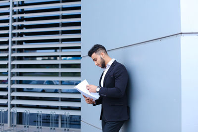 Young man reading document while standing by wall outdoors