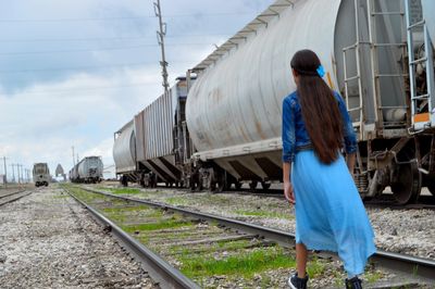 Rear view of woman wearing blue dress walking on railroad track by freight train against cloudy sky
