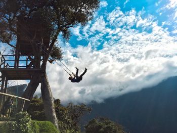 Woman standing by man swinging against cloudy sky