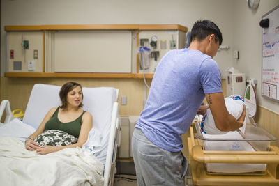 A father rests his newborn in a hospital crib while his wife looks on