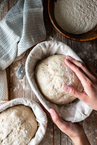 Woman hands from above holding sourdough bread on wicker stand with cloth on wooden table with flour scattered