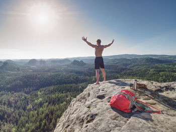 Shirtless sportsman take off backpack and rope on top of mountain and raised arms to greeting nature