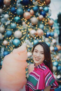 Portrait of smiling young woman standing by christmas tree holding cotton candy outdoors